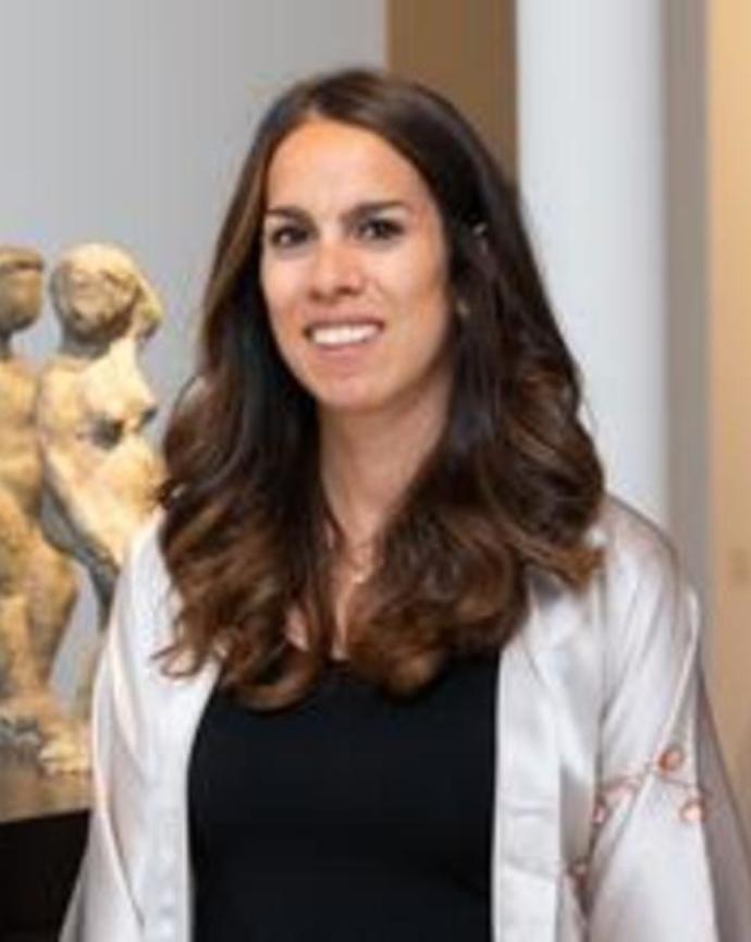 Portrait of a woman with long wavy hair, wearing a black top and light jacket, standing indoors near a sculpture.